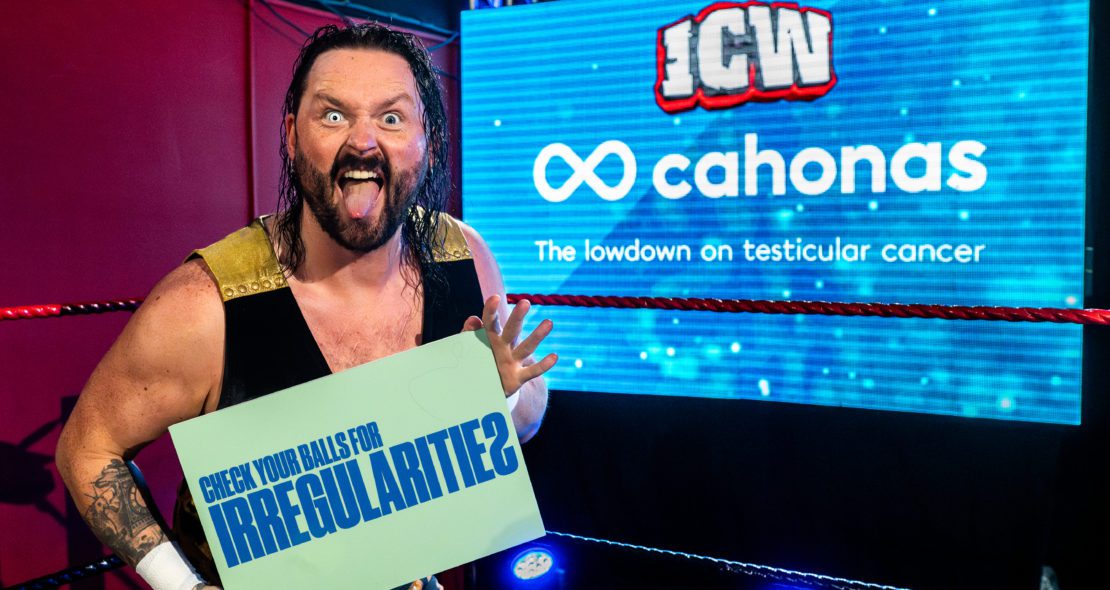 Wrestler holding testicular cancer awareness card which reads "Check for irregularities". This is attached to the "Wrestlers Slam Testicular Cancer Stigma" article.